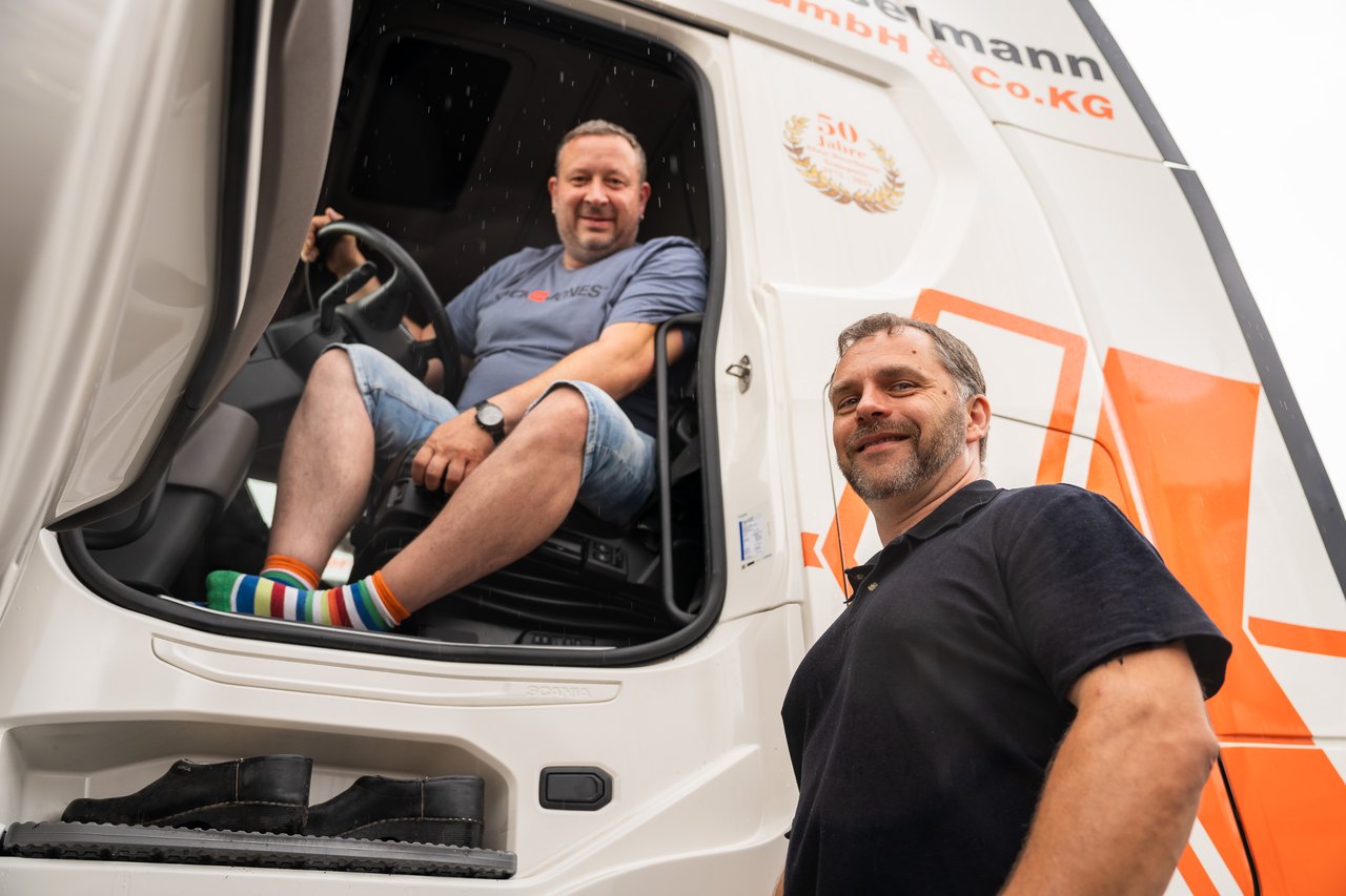 Zwei Männer lächeln; einer sitzt am Steuer eines weißen Lastwagens mit orangefarbenem Logo, der andere steht davor.